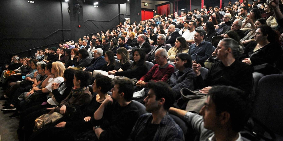 Modern bir tiyatro salonunda sahneye odaklanmış, dolu bir izleyici kitlesi. Farklı yaş gruplarından insanlar, koyu renkli koltuklarda oturarak dikkatle izledikleri gösteriyi takip ediyor. Salonun duvarları koyu gri renkte olup, arka planda kırmızı bir kapı ve acil çıkış tabelası görülüyor.
