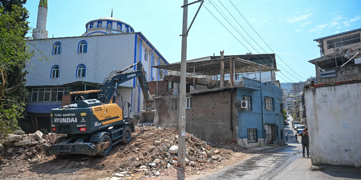 Altındağ'da bir sokak üzerinde, sarı ve siyah renkli Hyundai marka bir iş makinesi eski bir binanın yıkımını gerçekleştiriyor. Yıkım alanının hemen yanında beyaz ve mavi detaylı bir cami ile mavi boyalı eski bir ev bulunuyor. Sokak üzerinde bir kişi yürürken, arka planda diğer binalar ve trafik akışı görülüyor.