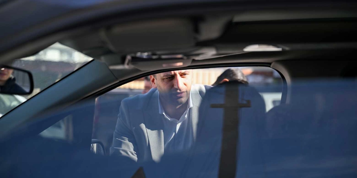 Açık renk ceket giymiş bir erkek, park halindeki bir otomobilin açık olan sürücü kapısından içeriye doğru eğilmiş, sürücü koltuğundaki kişiyle konuşuyor.