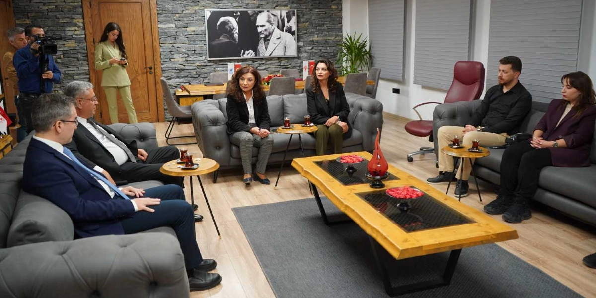 Modern bir ofis ortamında, karşılıklı gri koltuklarda oturan bir grup insan toplantı yapıyor. Ortada doğal ahşap görünümlü bir sehpa üzerinde çay bardakları ve kırmızı dekoratif objeler bulunuyor. Arka planda taş duvar üzerinde Mustafa Kemal Atatürk'ün siyah beyaz bir portresi asılı. Odanın köşesinde bir kameraman çekim yapıyor, diğer katılımcılar ise dikkatle dinliyor. Genel atmosfer resmi ve profesyonel bir çalışma toplantısını yansıtıyor.