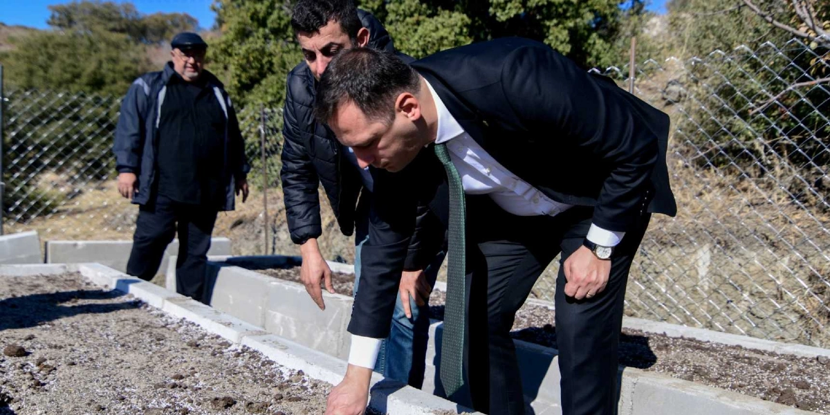 Dış mekanda, beton bloklarla çevrili toprak bir alanda, takım elbiseli ve kravatlı bir adam eğilerek toprağı inceliyor. Yanında ona eşlik eden iki kişi daha bulunuyor; biri koyu renkli bir ceket giymiş, diğeri ise arka planda duran daha yaşlı bir adam. Arka planda tel örgü ve ağaçlık bir arazi yer alıyor.