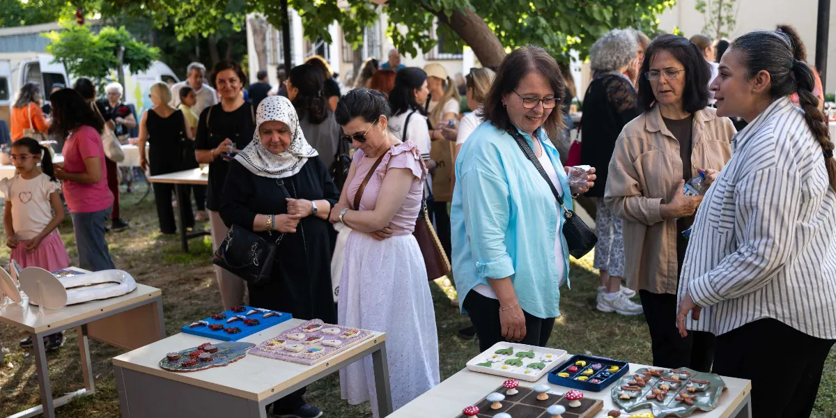 Ağaç gölgesindeki bir bahçede kurulan stantlarda sergilenen el yapımı seramik ve dekoratif objeleri inceleyen bir grup kadın. Ön planda, üzerinde minyatür mantar figürleri, renkli taşlar ve çeşitli süs eşyalarının bulunduğu masalar yer alıyor. Arka planda ise etkinliğe katılan diğer insanlar, masalar ve ağaçlar görülüyor.
