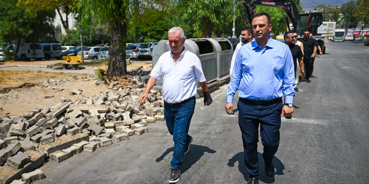 Bornova merkezindeki bir cadde üzerinde, beyaz gömlekli ve mavi kot pantolonlu bir erkek ile açık mavi gömlekli ve lacivert pantolonlu bir erkeğin yan yana yürüdüğü görülüyor. Arka planda, devam eden bir çevre düzenleme çalışması kapsamında sökülmüş parke taşları, ağaçlar, park halindeki araçlar ve bir iş makinesi yer alıyor. İki erkeğin arkasında ise onları takip eden birkaç kişi daha bulunuyor.
