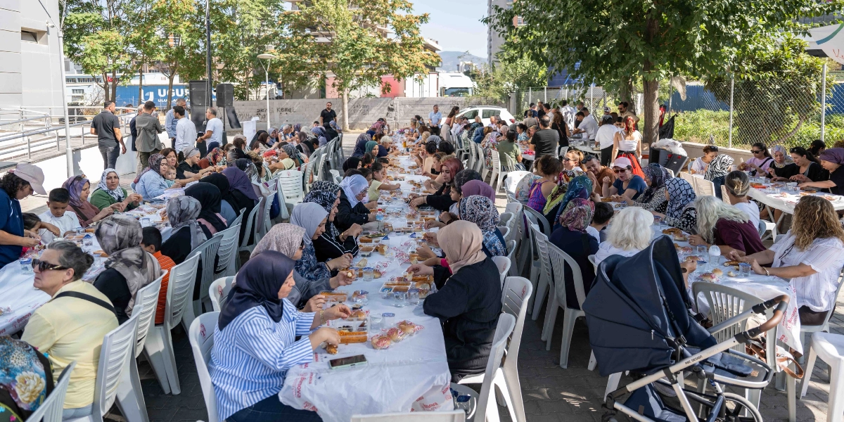Altındağ Kent Bostanı'nda, beyaz örtülü uzun masaların etrafında toplanmış çok sayıda kişinin katıldığı bir menemen etkinliği. İnsanlar yan yana dizilmiş plastik sandalyelerde oturarak yemek yiyor ve sohbet ediyor. Açık havada, ağaçların gölgesinde gerçekleşen etkinlikte, masaların üzerinde tek kullanımlık kaplarda menemen ve ekmekler bulunuyor. Ön planda bir bebek arabası ve çeşitli kıyafetler içindeki katılımcılar görülüyor.