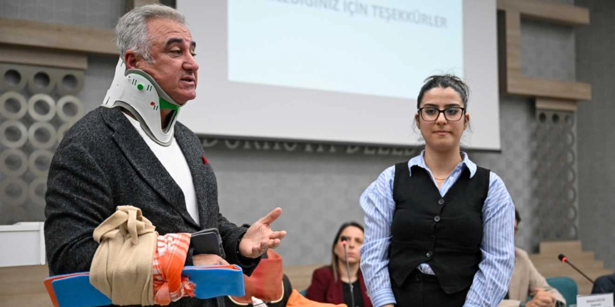 Boynunda tıbbi boyunluk bulunan ve kolu askıda olan orta yaşlı bir erkek, bir toplantı salonunda konuşma yaparken yanında genç bir kadın duruyor. Arka planda üzerinde 