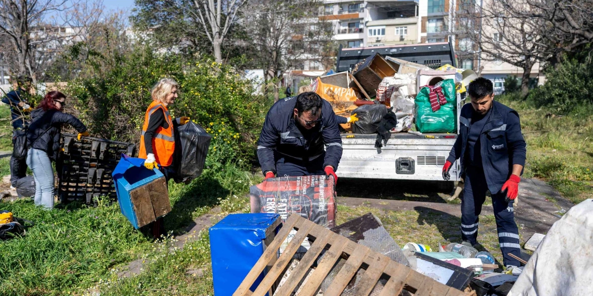 Belediye görevlileri ve gönüllüler, park alanındaki atıkları ve eski eşyaları toplayarak arkası dolu bir kamyonete yüklüyor. Çalışanlar eldiven takmış durumda ve çevredeki plastik kutular, ahşap paletler ve çöp poşetleri gibi malzemeleri temizleyerek alanı düzenliyor.
