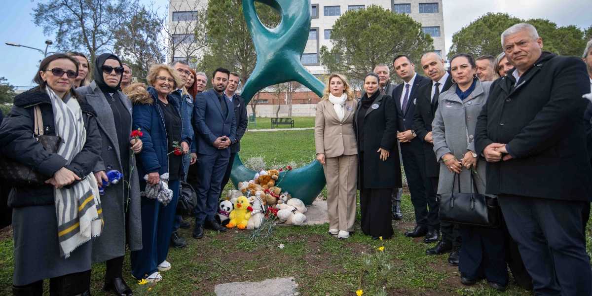Bornova'da yeşil bir heykelin önünde toplanmış, ellerinde karanfiller tutan bir grup insan, Türkan Bebek anması için bir araya gelmiş. Heykelin dibine oyuncak ayılar, peluş hayvanlar ve çiçekler bırakılmış. Katılımcılar resmi kıyafetler içinde, hüzünlü ve ciddi bir ifadeyle yan yana duruyor. Arka planda ise ağaçlar ve modern bir bina yapısı görülüyor.