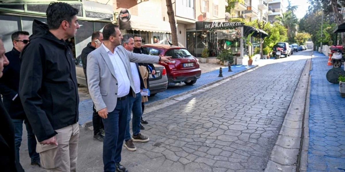 Gri ceketli bir yetkili, yanındaki bir grup insanla birlikte Bornova'da yenilenmiş, taş döşeli bir sokağı ve mavi renge boyanmış kaldırımları inceliyor; yetkili eliyle sokağın ilerisini işaret ederek yanındakilere bilgi veriyor.