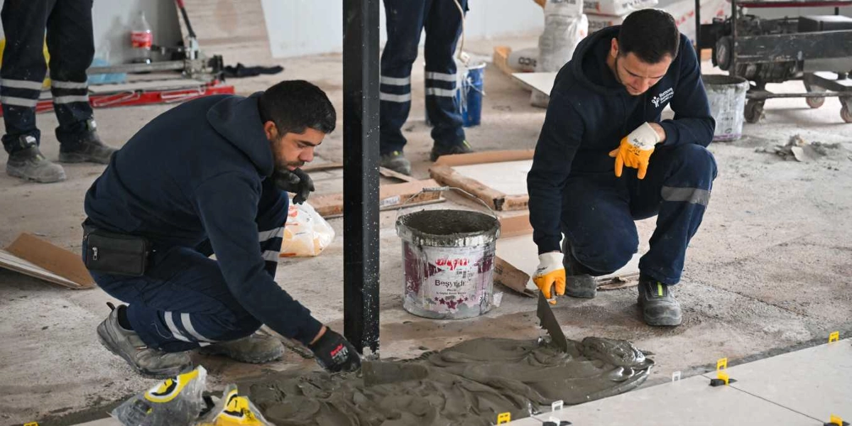 İnşaat halindeki bir alanda, lacivert iş kıyafetleri giymiş iki işçi yerde fayans döşeme çalışması yapıyor. İşçilerden biri elindeki mala ile harcı zemine yayarken, diğeri fayansları yerleştiriyor. Aralarında harç dolu bir kova bulunuyor ve arka planda inşaat malzemeleri ile diğer çalışanların ayakları görünüyor.