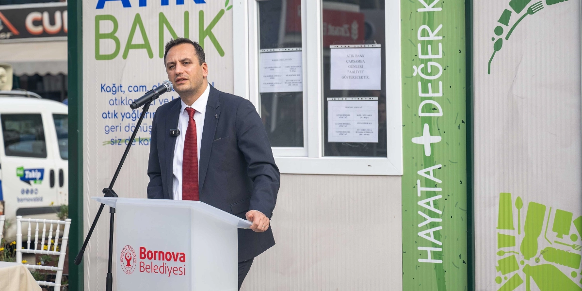 Koyu renk takım elbiseli ve kırmızı kravatlı bir erkek, üzerinde Bornova Belediyesi logosu bulunan bir kürsüde mikrofon başında konuşma yapıyor. Konuşmacının arkasında, üzerinde 