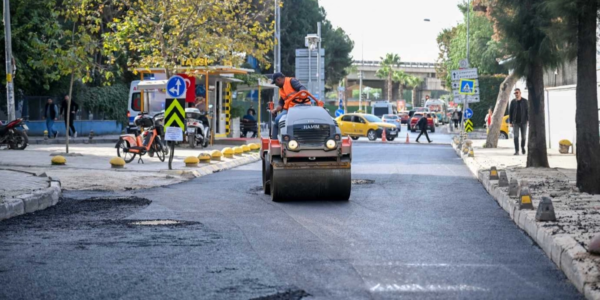 Süvari Caddesi'nde devam eden yol yenileme çalışmaları kapsamında, turuncu yelekli bir operatörün kullandığı HAMM marka silindir, yeni serilmiş asfalt üzerinde ilerliyor. Arka planda trafik akışı devam ederken, yol kenarında bir taksi durağı, trafik işaretleri ve ağaçlar bulunuyor.