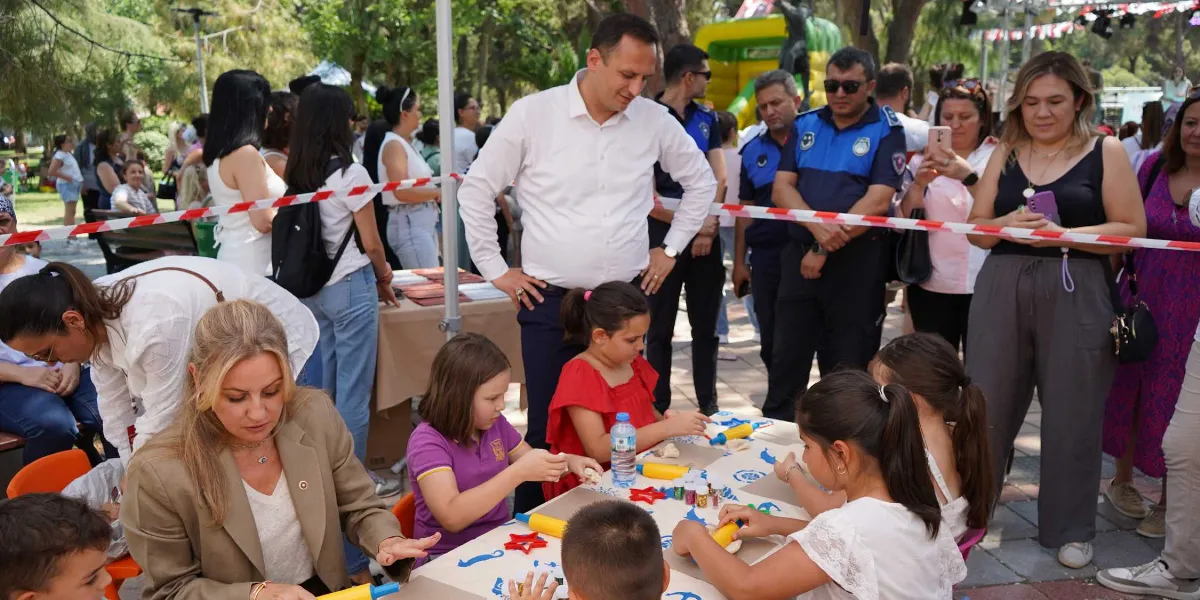 Açık havada düzenlenen çocuk şenliğinde, bir masanın etrafında toplanmış çocuklar oyun hamurlarıyla etkinlik yapıyor. Masanın başında sarı ceketli bir kadın çocuklara eşlik ederken, arkalarında beyaz gömlekli bir erkek, üniformalı görevliler ve etkinliği izleyen yetişkinler bulunuyor. Arka planda ağaçlık bir alan ve çocuklar için kurulmuş şişme oyun parkı görülüyor.