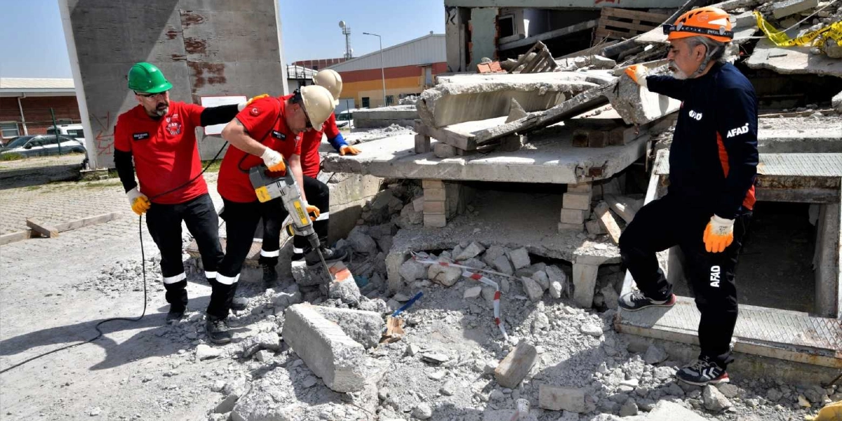 AFAD ekipleri, yıkılmış bir binanın enkaz alanında arama kurtarma çalışması yürütüyor. Kırmızı üniformalı ve baretli iki görevli, ellerindeki kırıcı delici aletle beton parçalarını kırmaya çalışırken, yanlarında duran başka bir görevli ise çalışmaları gözlemliyor. Ekip üyeleri iş güvenliği eldivenleri ve koruyucu ekipmanlar kullanıyor.