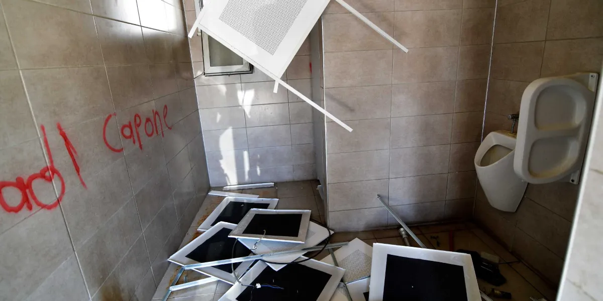 Tahrip edilmiş bir umumi tuvalette, yerlere saçılmış tavan panelleri ve duvarda kırmızı sprey boyayla yazılmış 