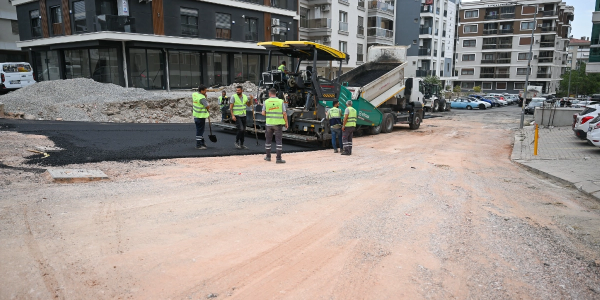 Şehir merkezindeki bir sokakta, sarı ikaz yelekli işçiler asfalt serme makinesi ve kamyon kullanarak yol yapım çalışması yürütüyor. İşçiler, yeni dökülen sıcak asfaltın üzerinde ellerinde küreklerle çalışırken, arka planda modern apartmanlar ve park halindeki araçlar görülüyor.