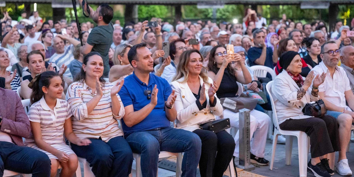 Açık havada düzenlenen bir etkinlikte, beyaz plastik sandalyelerde yan yana oturan ve alkışlayarak gösteriyi izleyen kalabalık bir seyirci grubu. Ön planda, çizgili tişörtlü bir kız çocuğu, yanında çizgili gömlekli bir kadın, mavi polo yaka tişörtlü bir erkek ve beyaz ceketli bir kadın yer alıyor. Arka planda ise cep telefonlarıyla çekim yapan veya sahneyi izleyen çok sayıda kişi bulunuyor.