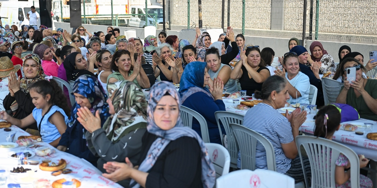 Açık havada, uzun masaların etrafında toplanmış, çoğunluğu başörtülü kadınlardan oluşan kalabalık bir grup, neşeyle alkış tutuyor. Masaların üzerinde Bornova Belediyesi logolu örtüler, simitler ve çeşitli yiyecekler bulunuyor. Bazı kadınlar gülümseyerek ellerini çırparken, bazıları da o anı cep telefonlarıyla kaydediyor. Ortamda samimi ve kutlama havası hakim.