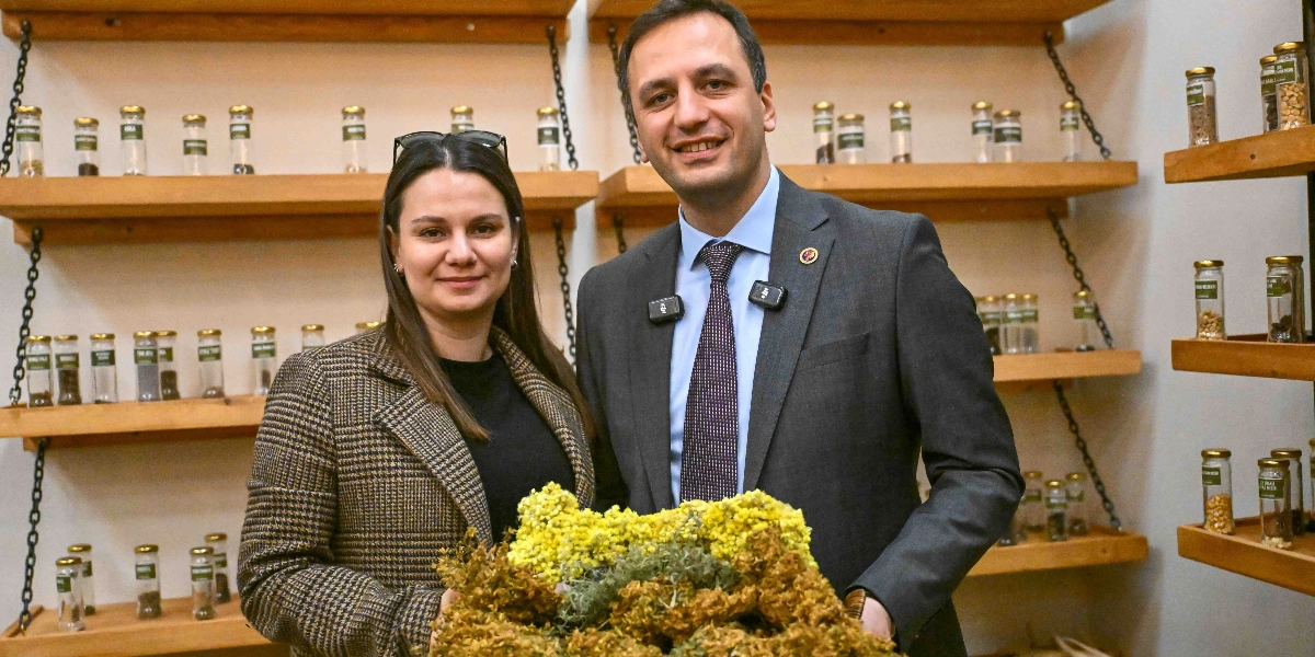 Koyu renkli ekose bir ceket giyen bir kadın ve lacivert takım elbiseli, kravatlı bir erkek, arkalarında raflara dizilmiş çok sayıda küçük cam kavanoz içindeki tohumların bulunduğu bir ortamda, ellerinde tuttukları sarı çiçekli bitki demetiyle yan yana durarak gülümsüyor.