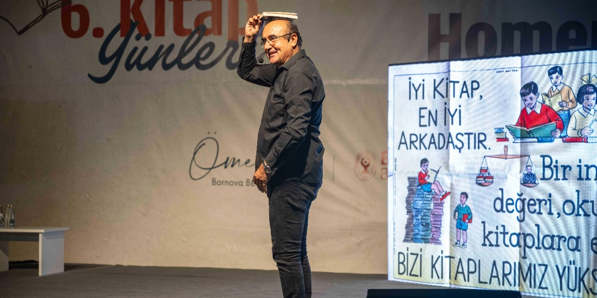 Siyah gömlek ve pantolon giyen gözlüklü bir erkek, sahnede başının üzerinde bir kitap dengede dururken gülümsüyor. Arka planda 