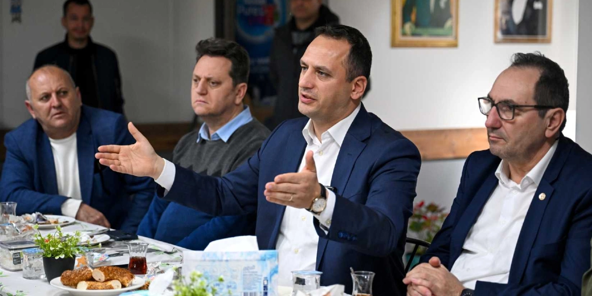 Bir toplantı masasında oturan dört erkekten merkezde bulunan kişi, elleriyle jest yaparak konuşma yapıyor. Masada çay bardakları, bir tabak simit ve peçetelik bulunuyor. Katılımcılar resmi kıyafetler içinde, dikkatle dinliyorlar.