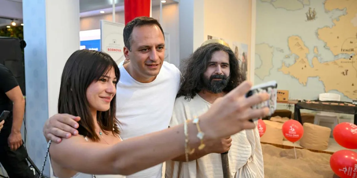 Bornova Belediye Başkanı Ömer Eşmi, fuar standında genç bir kadın ve Homeros kostümü giymiş uzun saçlı, sakallı bir oyuncu ile birlikte selfie çekiliyor. Arka planda İzmir haritasının yer aldığı bir duvar dekoru ve üzerinde Bornova Belediyesi logoları bulunan kırmızı balonlar görülüyor.