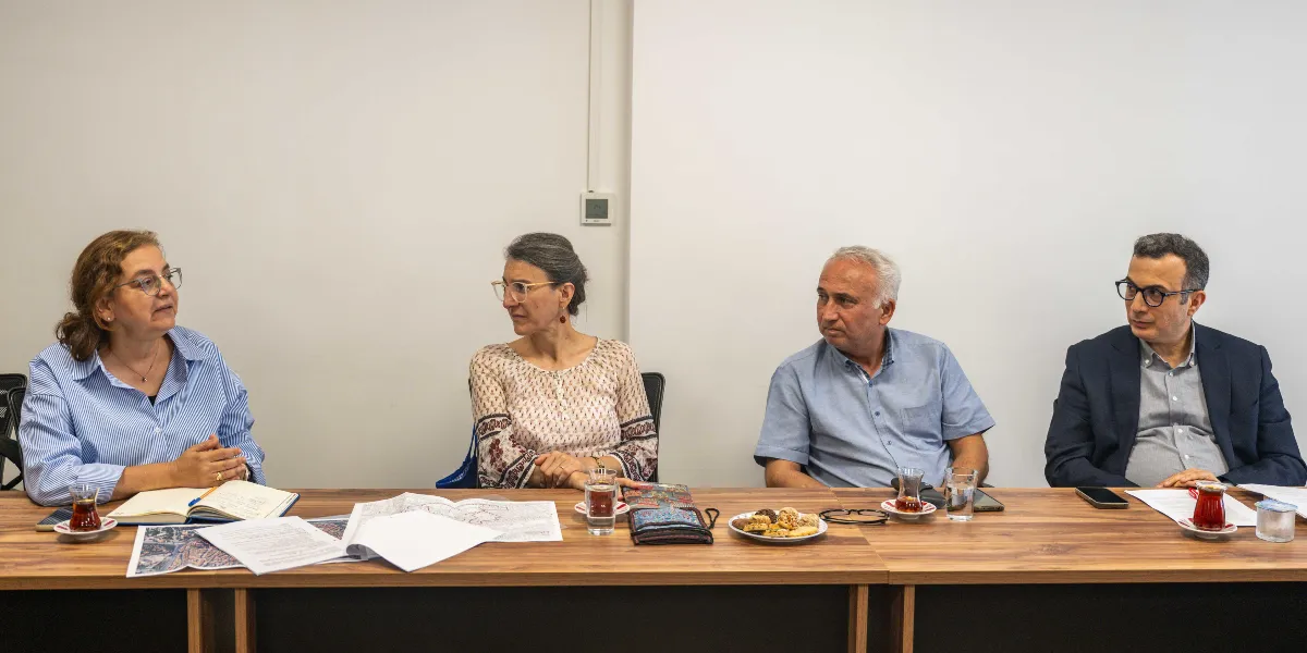 Beyaz bir duvarın önünde, ahşap bir toplantı masasında yan yana oturan dört kişi bir toplantı gerçekleştiriyor. Soldan sağa doğru; mavi beyaz çizgili gömlekli gözlüklü bir kadın, desenli bluz giyen gözlüklü bir kadın, kısa kollu mavi gömlekli bir erkek ve lacivert ceketli gözlüklü bir erkek yer alıyor. Masanın üzerinde haritalar, not defterleri, kalemler, bir tabak kurabiye ve ince belli bardaklarda çaylar bulunuyor.