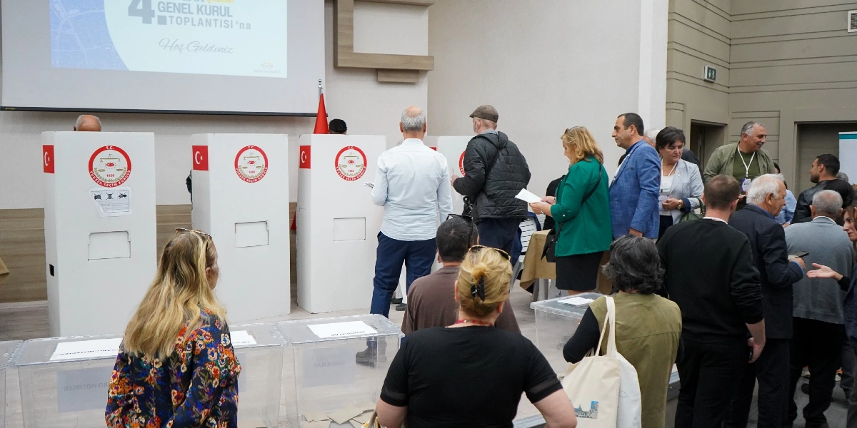 Kapalı bir salonda düzenlenen seçimde, üzerinde T.C. Yüksek Seçim Kurulu amblemi bulunan beyaz oy verme kabinlerinin önünde sıraya girmiş insanlar ve önlerinde şeffaf oy sandıkları görülüyor. Arka planda duvardaki projeksiyon ekranında 4. Genel Kurul Toplantısı'na Hoş Geldiniz yazısı yer alıyor.