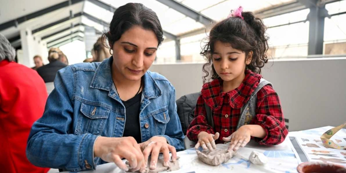 Kot ceket giyen bir kadın ve kırmızı ekose gömlekli küçük bir kız çocuğu, bir masanın başında oturmuş dikkatle kil hamuruna şekil veriyor. Arka planda başka insanların da bulunduğu aydınlık bir atölye ortamında, ikili birlikte seramik çalışması yapıyor.