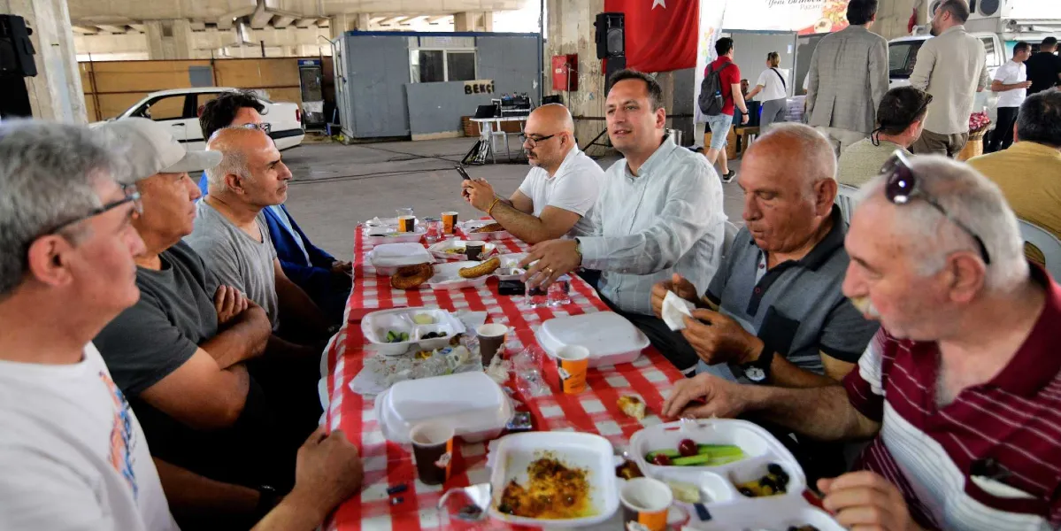 Kapalı bir alanda, kırmızı beyaz kareli örtü serili uzun bir masanın etrafında toplanmış bir grup erkek, kahvaltı eşliğinde sohbet ediyor. Masanın üzerinde plastik tabaklarda zeytin, peynir, domates ve simit gibi yiyecekler ile karton bardaklarda içecekler bulunuyor. Gruptakilerden biri konuşurken diğerleri onu dinliyor. Arka planda Türk bayrağı, park halindeki bir otomobil ve ayakta duran başka insanlar yer alıyor.