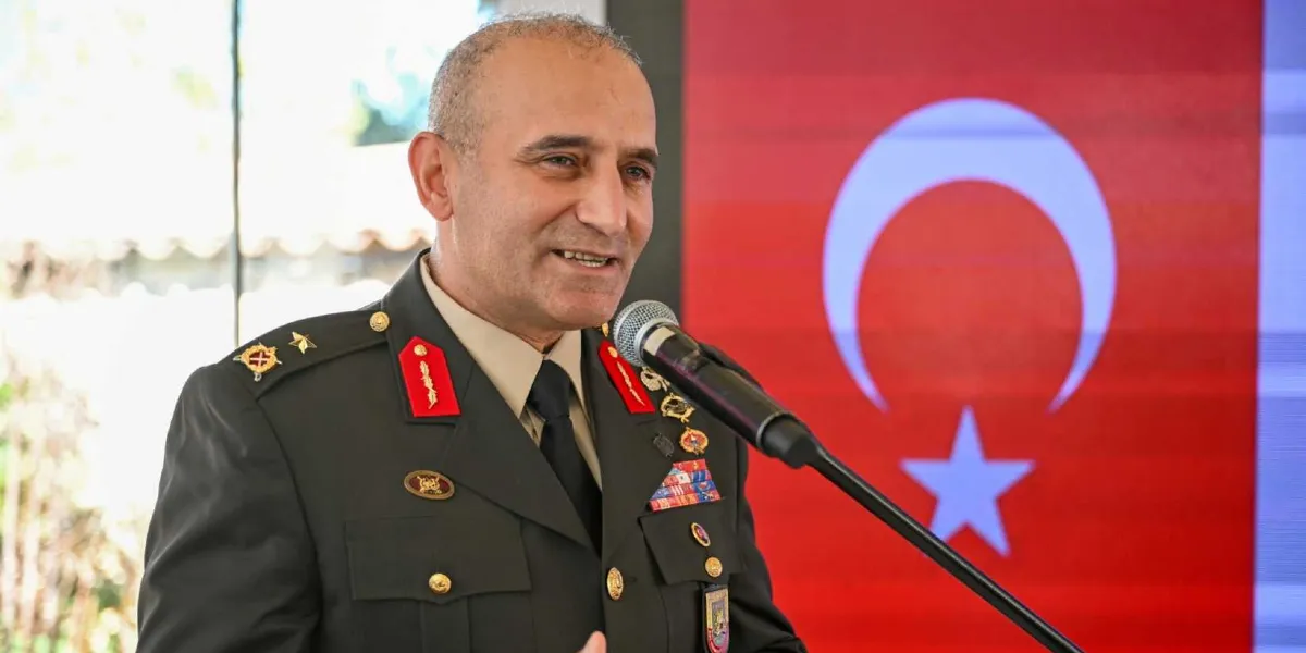 Resmi üniformalı, rütbeli bir asker, önünde duran mikrofona konuşma yapıyor. Arka planda büyük bir Türk bayrağı görseli yer alıyor.