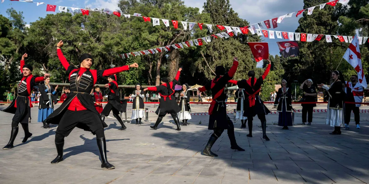 Açık havada, üzerinde Türk bayrakları ve Atatürk posterlerinin asılı olduğu süslemelerin altında, geleneksel Kafkas kıyafetleri giymiş bir grup dansçı halk oyunu sergiliyor. Siyah ve kırmızı renkli, göğüs kısmında fişeklik detayları bulunan kostümleriyle erkek dansçılar ön planda yer alırken, arka planda yöresel kıyafetli kadın dansçılar ve izleyiciler bulunuyor. Sağ tarafta Gürcistan bayrağı tutan bir kişi dikkat çekiyor.