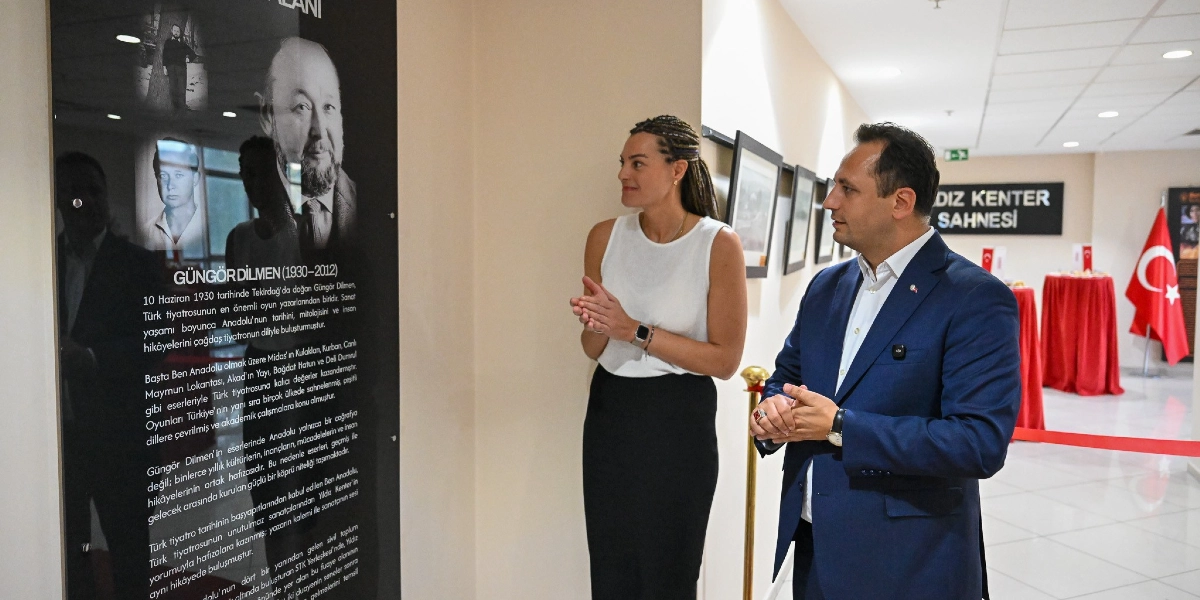Beyaz kolsuz bluz ve siyah etek giymiş bir kadın ile lacivert ceketli bir erkek, üzerinde Güngör Dilmen'in fotoğrafları ve biyografisinin yer aldığı siyah bir panoyu inceliyor. Arka planda Yıldız Kenter Sahnesi yazılı bir tabela ve Türk bayrağı görülüyor.