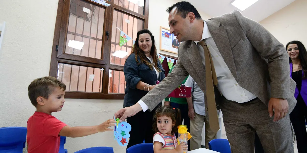 Bej renkli takım elbiseli bir erkek, sınıfta oturan küçük bir çocuğa üzerinde yıldız süslemeleri olan kelebek şeklinde bir kart hediye ediyor. Çocuğun yanında başka bir kız çocuğu ve arka planda gülümseyerek izleyen iki kadın bulunuyor. Sınıf ortamında, pencerelerde süslemeler ve duvarda Türk bayrağı ile Atatürk portresi yer alıyor.