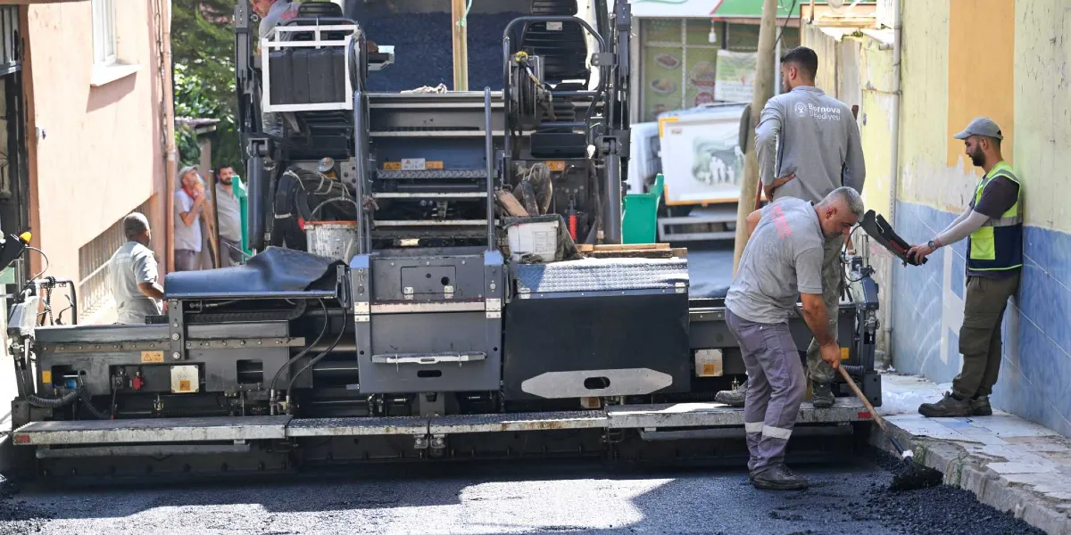 Dar bir sokakta asfalt serme makinesiyle yol çalışması yapan Bornova Belediyesi personeli. İşçilerden biri elindeki süpürgeyle asfaltı düzeltirken, diğerleri makinenin başında ve yanında duruyor. Personelin üzerinde Bornova Belediyesi logolu gri iş kıyafetleri ve yelekler bulunuyor.