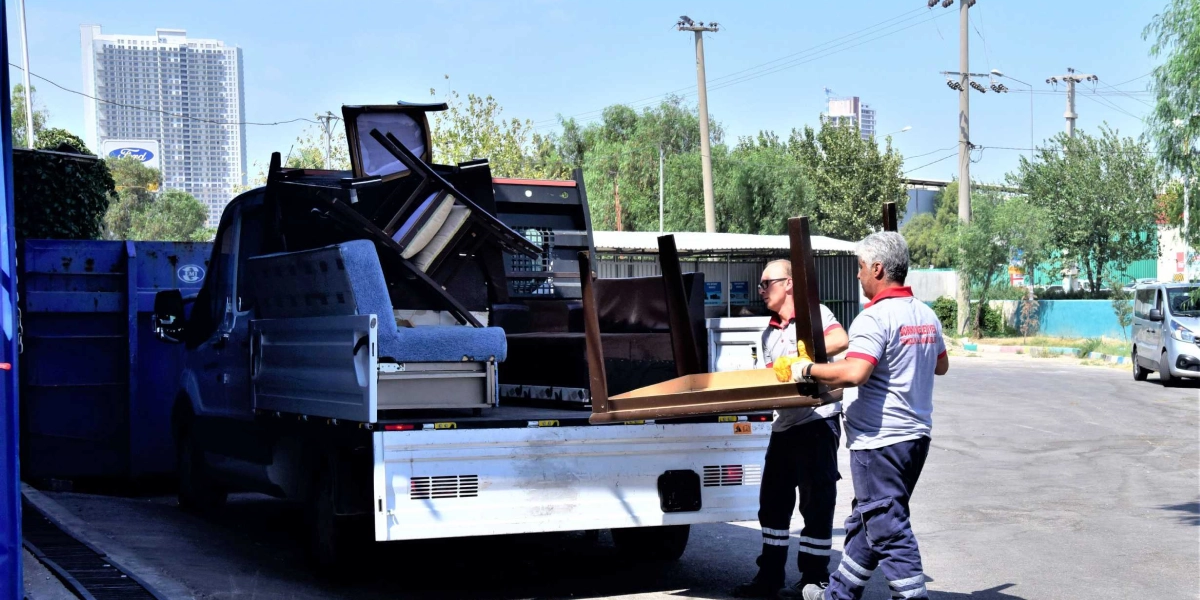 Bornova Belediyesi üniformalı iki görevli, selden zarar görmüş eşyalarla dolu bir kamyonetin kasasına ahşap bir masa yerleştiriyor. Kamyonetin kasasında koltuk ve çeşitli mobilya parçaları bulunuyor.