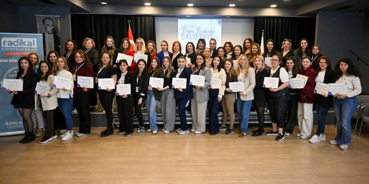 Bir konferans salonunda, ellerinde katılım belgeleriyle yan yana dizilmiş, çoğunluğu kadınlardan oluşan kalabalık bir grup öğretmen veya eğitimci toplu fotoğraf çektiriyor. Arka planda 