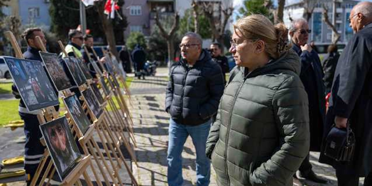 Açık havada, ahşap şövaleler üzerinde sergilenen Hocalı Katliamı'nı anlatan fotoğrafları inceleyen bir grup insan. Ön planda haki renkli şişme mont giymiş bir kadın ve yanında gözlüklü bir adam sergiyi dikkatle izliyor. Arka planda ise diğer katılımcılar ve ağaçlıklı bir park alanı görülüyor.