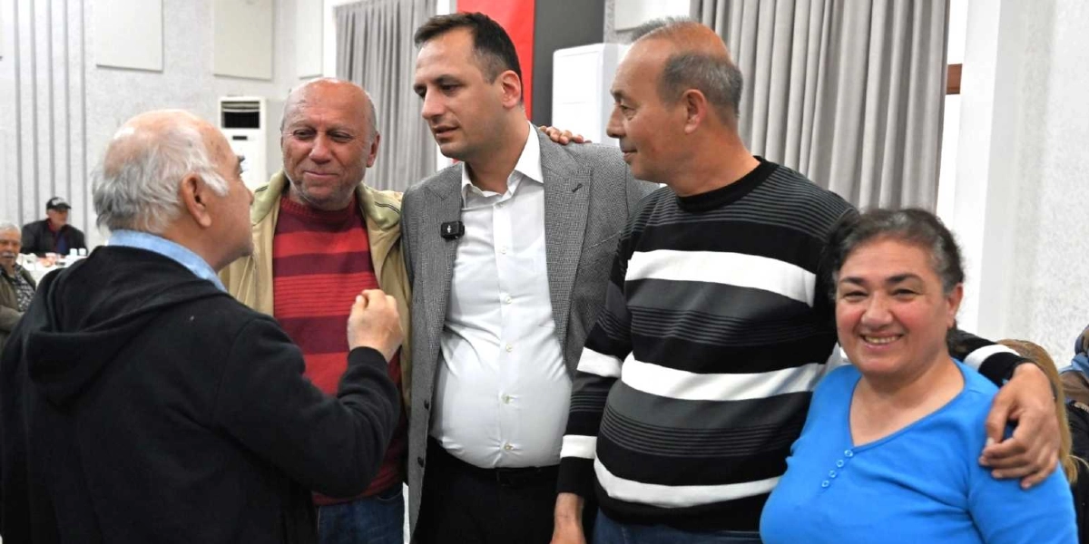 Kapalı bir mekanda düzenlenen dayanışma yemeğinde, yan yana duran beş kişilik bir grup samimi bir şekilde sohbet ediyor. Grubun solunda, sırtı kameraya dönük olan beyaz saçlı bir adam el hareketleriyle konuşurken, karşısında onu dinleyen kırmızı çizgili kazaklı ve bej ceketli bir adam, gri ceketli ve beyaz gömlekli bir bey, siyah beyaz çizgili kazaklı bir adam ve mavi bluzlu gülen bir kadın yer alıyor. Arka planda gri perdeler ve diğer masalarda oturan insanlar görünüyor.