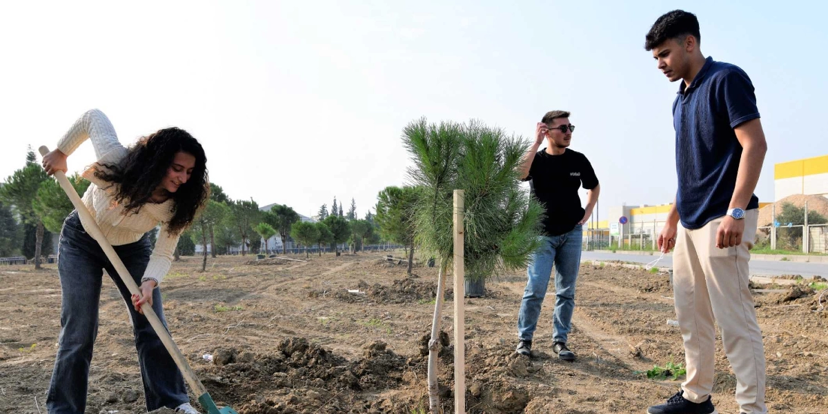 Açık bir alanda, toprak zeminde fidan diken üç genç. Sol tarafta beyaz kazaklı ve kot pantolonlu genç bir kadın elinde kürekle toprağı kazarken, sağ tarafta siyah tişörtlü ve güneş gözlüklü bir genç ile lacivert polo yaka tişörtlü başka bir genç onu izliyor. Arka planda yeni dikilmiş küçük çam ağaçları ve uzakta bazı yapılar görülüyor.