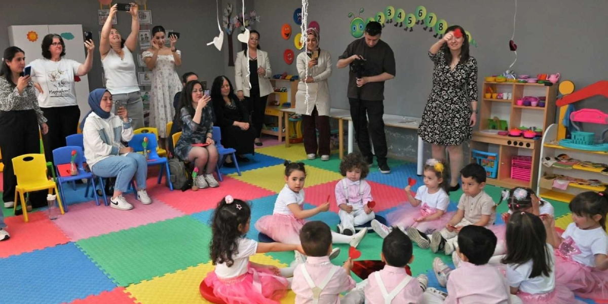 Bir anaokulu sınıfında Anneler Günü kutlaması için toplanmış çocuklar ve onları izleyen yetişkinler. Renkli yer minderleri üzerinde oturan çocuklar, beyaz tişörtler ve pembe tüller giymiş kızlar ile açık renkli gömlekler giymiş erkeklerden oluşuyor. Çocuklar ellerinde küçük kırmızı kalp figürleri tutuyor. Arka planda ayakta duran anneler ve öğretmenler, cep telefonlarıyla çocukların fotoğraflarını çekiyor. Sınıfın duvarında sayıların olduğu bir görsel ve köşede oyuncak mutfak setleri bulunuyor.