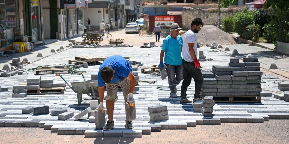 Bornova'da bir sokak yenileme çalışması kapsamında üç işçi, gri ve beyaz kilitli parke taşlarını döşeyerek yolu düzenliyor. Ön planda mavi tişörtlü bir işçi eğilmiş vaziyette taşları yerleştirirken, arkasında şapkalı ve mavi tişörtlü bir işçi ile beyaz tişörtlü bir başka işçi çalışmaya devam ediyor. Arka planda ise üzerinde 
