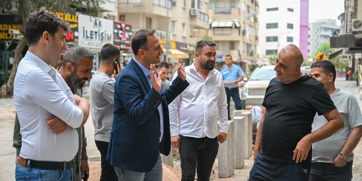 Bornova Zafer Caddesi'nde bir grup erkek, devam eden çevre düzenleme çalışmalarını inceliyor. Lacivert ceketli bir kişi ellerini havaya kaldırarak bir şeyler anlatırken, çevresindeki diğer kişiler onu dinliyor. Arka planda dükkan tabelaları, park halindeki araçlar ve yol kenarına yerleştirilmiş beton bariyerler görülüyor.