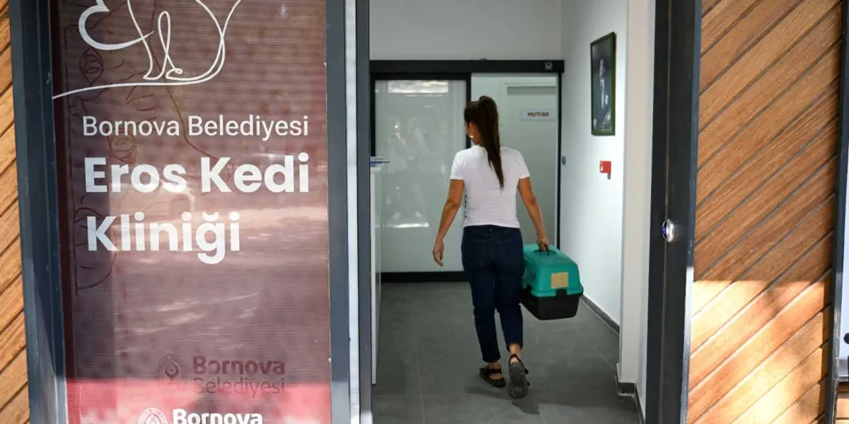Beyaz tişörtlü ve kot pantolonlu bir kadın, elinde yeşil bir kedi taşıma çantasıyla Bornova Belediyesi Eros Kedi Kliniği'nin girişinden içeriye doğru yürüyor. Kliniğin cam kapısında Bornova Belediyesi logosu ve Eros Kedi Kliniği yazısı yer alıyor.