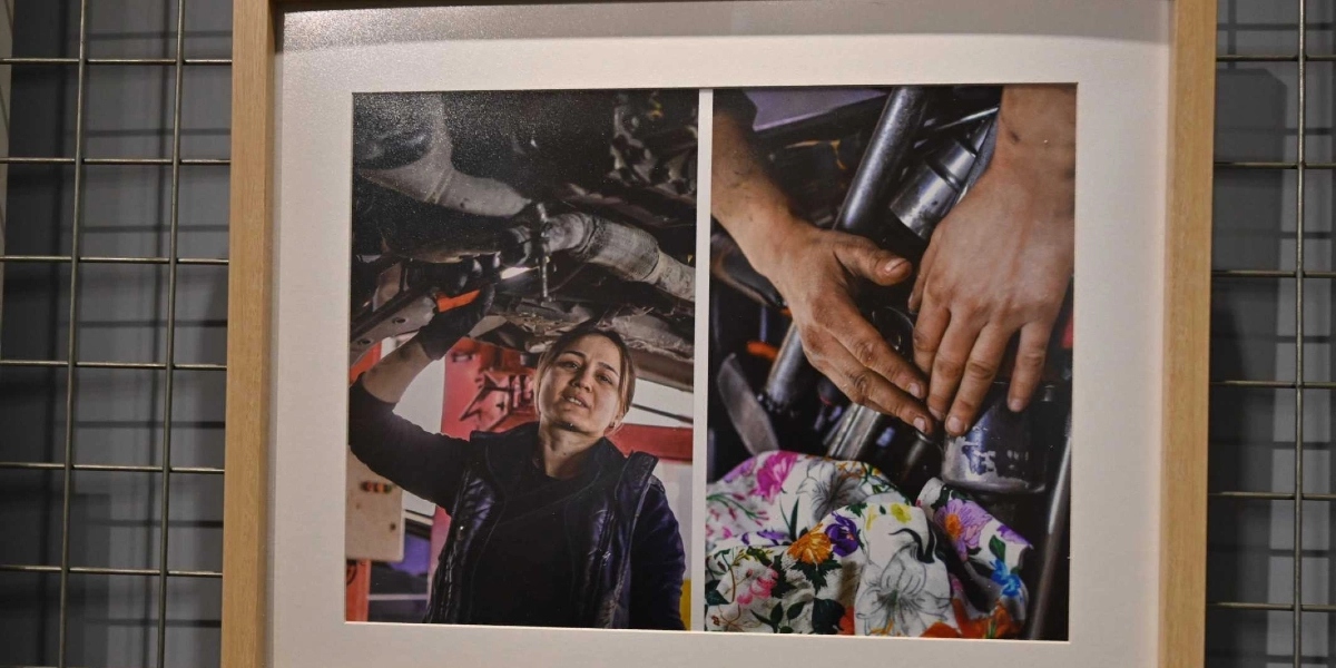 Ahşap çerçeve içinde yan yana yerleştirilmiş iki fotoğraftan oluşan bir sergi panosu. Sol taraftaki fotoğrafta, bir otomobilin altında tamir yapan, üzerinde siyah yelek bulunan bir kadın yer alıyor. Sağ taraftaki fotoğrafta ise, çiçek desenli bir kumaşın üzerinde duran ve bir motor parçasıyla uğraşan bir kişinin elleri görülüyor.