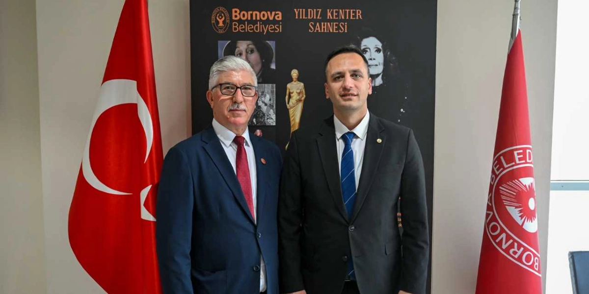 Arka planda Bornova Belediyesi Yıldız Kenter Sahnesi yazılı bir pano önünde, yan yana duran iki erkek. Soldaki kişi gri saçlı, gözlüklü, lacivert takım elbiseli ve kırmızı kravatlı; sağdaki kişi ise kısa saçlı, lacivert takım elbiseli ve mavi kravatlıdır. İkilinin sağında ve solunda Türk bayrağı ile Bornova Belediyesi bayrağı yer almaktadır.