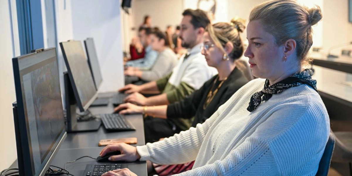 Aydınlık bir ofis ortamında yan yana dizilmiş bilgisayar masalarında çalışan bir grup insan. Ön planda, beyaz kazaklı ve saçları topuz yapılmış bir kadın, bilgisayar başında dikkatle çalışıyor. Onun hemen arkasında diğer çalışanlar da kendi bilgisayarlarında benzer şekilde odaklanmış durumda. Masaların üzerinde siyah klavyeler ve fareler bulunuyor.