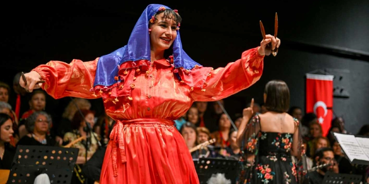 Geleneksel kırmızı kıyafetli ve mavi başörtülü bir kadın, ellerinde tuttuğu ahşap kaşıkları birbirine vurarak sahnede dans ediyor. Kadının arkasında, orkestra düzeninde oturan müzisyenler ve bir koro grubu yer alıyor. Arka planda ise Türk bayrağı görülüyor.