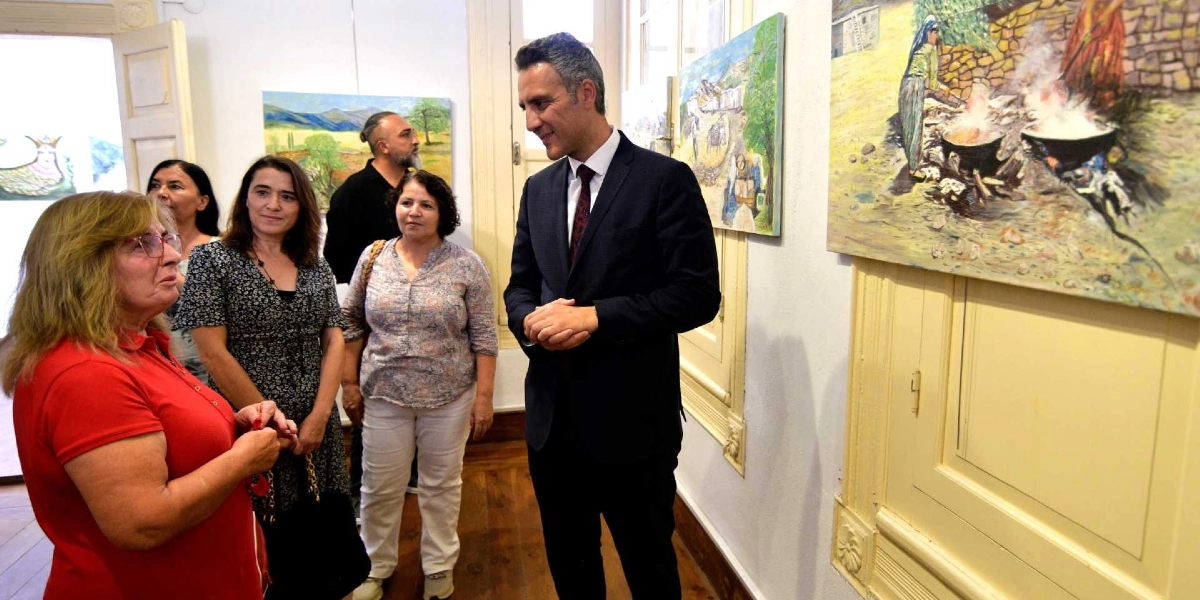 Dramalılar Köşkü'ndeki sergi salonunda, takım elbiseli bir erkek ve dört kadın, duvarda asılı olan Anadolu temalı yağlı boya tabloları inceliyor. Sağ taraftaki tabloda, dış mekanda ateş üzerinde yemek pişiren yöresel kıyafetli kadınlar resmedilmiş. Gruptakiler birbirleriyle sohbet ederek eserleri gözlemliyor.