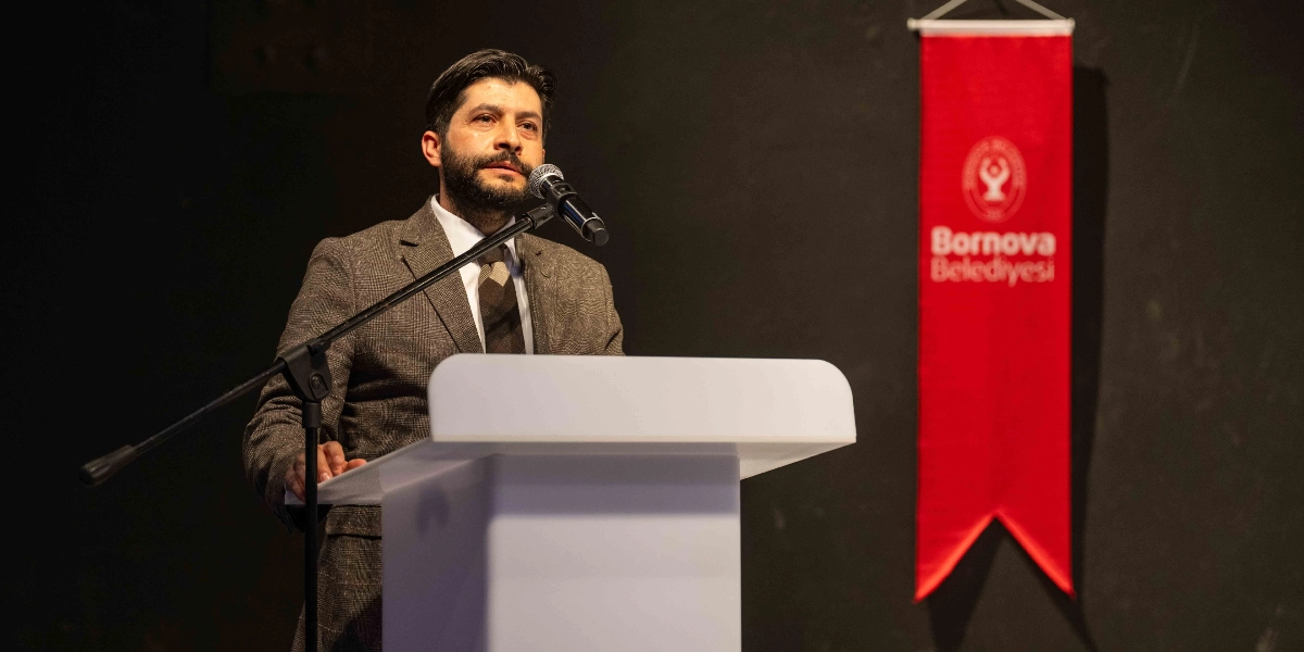 Koyu renkli bir arka plan önünde, beyaz bir kürsü arkasında duran, sakallı ve kahverengi ekose ceketli bir erkek konuşma yapıyor. Konuşmacının yanında, üzerinde Bornova Belediyesi logosu ve yazısı bulunan dikey kırmızı bir flama asılı duruyor.