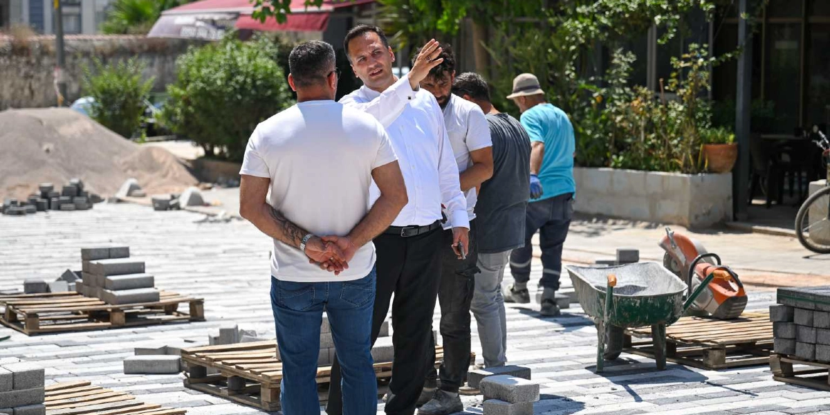 Beyaz gömlekli bir yetkili, devam eden bir çevre düzenleme çalışmasını yerinde inceleyerek elini ileriye doğru uzatıp yönlendirmelerde bulunuyor. Yanında duran ve sırtı dönük olan kişi ile diğer işçiler, döşenmeye hazır gri parke taşlarının bulunduğu şantiye alanında çalışmayı takip ediyor. Arka planda ise el arabası, kesim makinesi ve paletler üzerinde dizili taşlar yer alıyor.